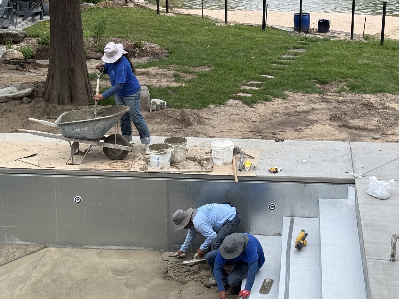 Crew hand-troweling the pool floor base, the lake visible just beyond the fence